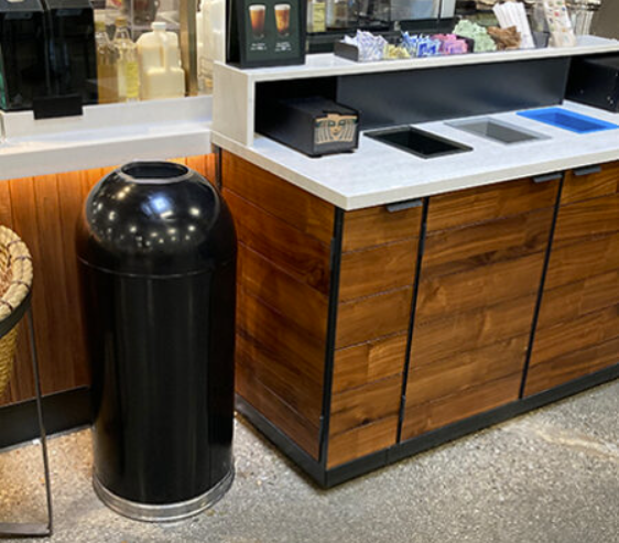 Black commercial indoor trash can placed beside a café beverage station, designed for high-traffic food service environments.